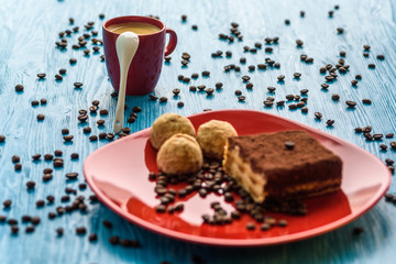 Tiramisu cake on a red plate, near a cup of coffee, photographed on a light wooden background.