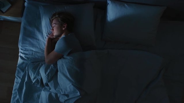 Top View of Handsome Young Man Sleeping Cozily on a Bed in His Bedroom at Night. Blue Nightly Colors with Cold Weak Lamppost Light Shining Through the Window.