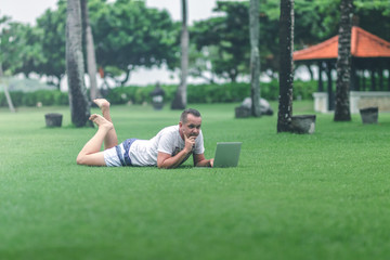 Naklejka premium Young man in the green tropical park working online with laptop. Freelancer man on Bali island concept.