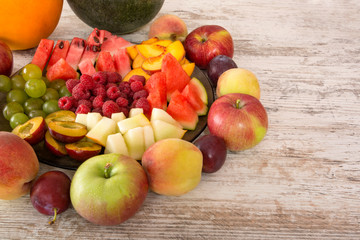 Fruit mix. Apple, peach, grapes, watermelon, cantaloupe, raspberry, plum. Whole fruits and slices on a plate lie on a white wooden background. Copy space, Place for text.