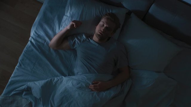 Top View of Handsome Young Man Sleeping Cozily on a Bed in His Bedroom at Night. Blue Nightly Colors with Cold Weak Lamppost Light Shining Through the Window.