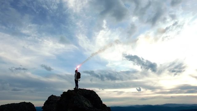 Young woman going to the top and burn the signal firecracker standing on the rock. Beautiful landscape with autumn forest and cloudy horizon at sunset. Successful wild journey, freedom and happiness