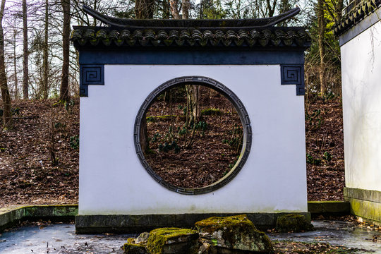 Brick Wall And Round Window Door Through To Open Chinese Garden And Small Bonsai Tree In A Pot.