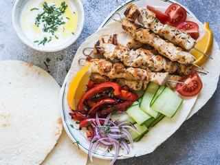 Grilled chicken on metal skewers with of fresh vegetables on the pita and Greek sauce in a bowl on a gray background. Greek cuisine.