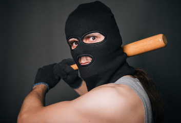 Bandit, the criminal in a black mask and in a gray T-shirt with a painless bat. Robber with a weapon. A man in a balaclava and a gray mask on a gray background