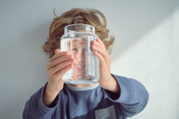 Cute little boy smiling and holding glass jar with clean water in front of face while standing near white wall