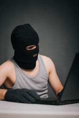 A thief in a black mask stole a laptop. A man in a balaclava and a computer in hand on a gray background. The hacker is hacking the computer. Steals information. A young man breaks the law.