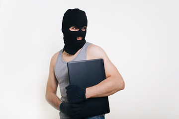 A thief in a black mask stole a laptop. A man in a balaclava and with a computer in hands on a white background. The hacker is hacking the computer. Steals information. A young man breaks the law.