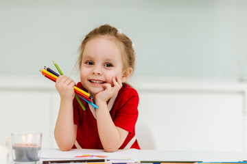 Cute little Caucasian girl in casual red dress draws and  sits at the table and holds colored pencils in his hands. Pozing amd making funny faces. Preparing a preschooler for school