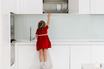 Cute smart little caucasian girl in casual red dress climbed onto a chair and trying to reach dishes in a closet in a modern minimalist kitchen