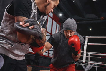 Close quarter boxing. Strong tattooed athlete in sports clothing training on boxing paws with...