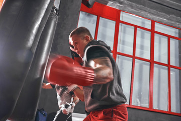 Ready to break his record. Young muscular athlete in sports clothing training hard on heavy punch bag before big fight in boxing gym