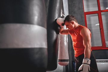 Tired after workout. Tired young athlete in sports clothing looking away after hard training on heavy punch bag in boxing gym