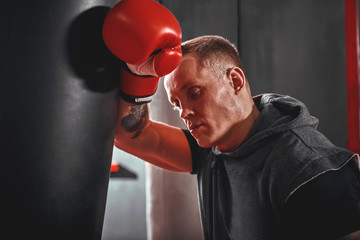 Tired after workout. Tired young athlete in sports clothing looking away after hard training on heavy punch bag in boxing gym