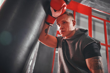 Taking time to rest. Tired young athlete in sports clothing taking break after hard training on heavy punch bag in boxing gym
