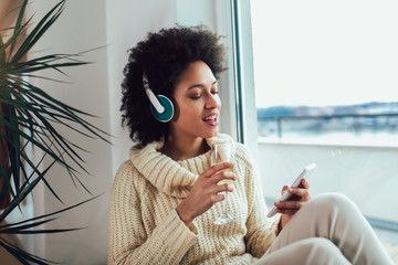 Young beautiful african american woman relaxing and listening to music using headphone, drinking wine.