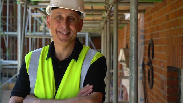 Male builder foreman, worker or architect working on construction building site standing on scaffolding folding his arms while laughing and smiling to camera