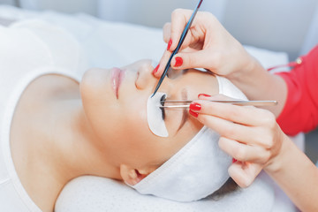 Eyelash Extension Procedure. Close up view of beautiful Woman with Long Eyelashes. Stylist holding tweezers, tongs and making lengthening lashes for girl in a beauty salon. Beauty Concept.
