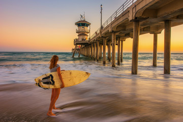 pier at sunset, young woman is watching the waves with a surboard