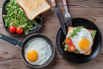 Top view of fried eggs on black plate and pan with fork, rukkola and mushrooms