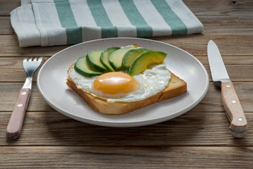 Toast bread with fried eggs, avocado. And cucumber slices on wooden table with fork and knife. Soft focus.