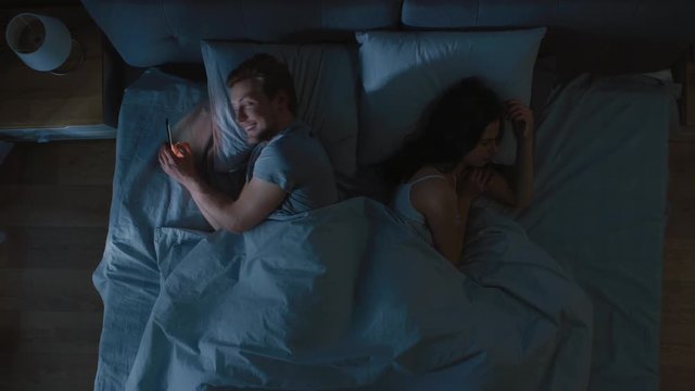 Cheating and Unfaithful Young Man Uses Smarphone for Finding a New Date or Bragging Night in Bed when Girlfriend Sleeps Soundly. Top View Camera