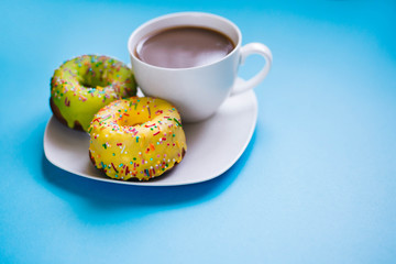 Two colored donuts on a plate with a cup of coffee. Yellow and Green Donut on a blue background. Dessert