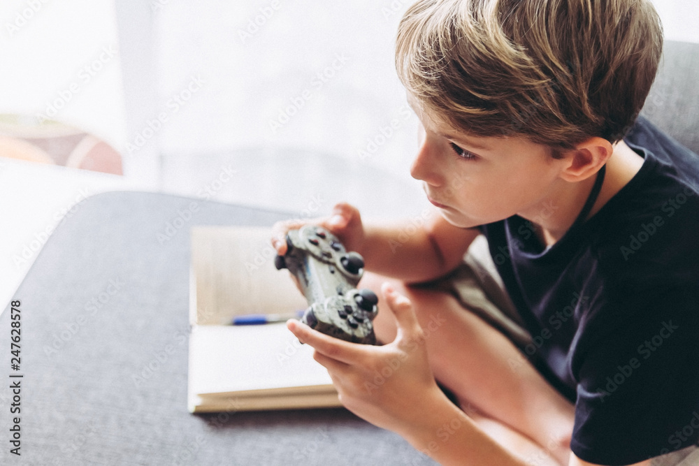 Wall mural Blond child playing video games instead of studying sitting home sofa with legs crossed young student resting playing online with friends domestic pastime challenge web Close up portrait white curtain