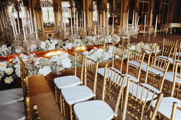 Decorations for wedding dinner. Golden vases with candles stand on white tables ready for fesrive reception