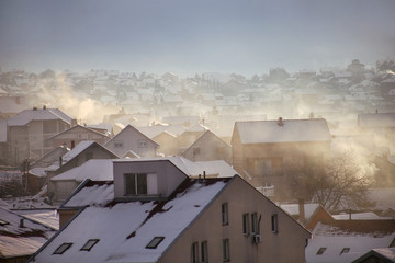 Smoking chimneys at roofs with snow of houses emits smoke, smog at sunrise, pollutants enter atmosphere. Environmental disaster. Harmful emissions, exhaust gases into air. Winter day, heating season.