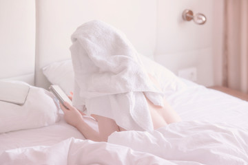 Fototapeta na wymiar Unrecognizable girl with a towel on her head in bed reading reading messages in smart phone, back view. Soft image