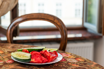 Chair by rustic vintage wooden antique table and window in Ukraine or Russia with plate of fresh sliced vegetables, cucumbers and tomatoes