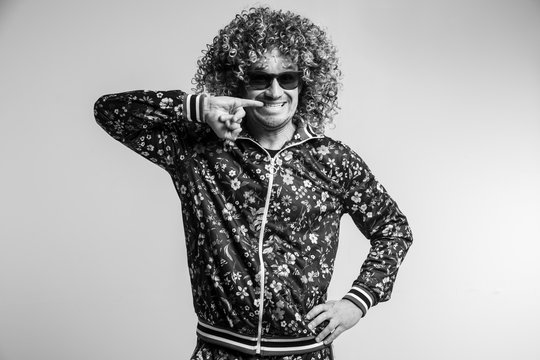 Indoor portrait of excited smiling man in fashionable trendy tracksuit with pattern and sunglasses touching teeth with finger. Adult cheerful male with curly hair fooling on white studio background.