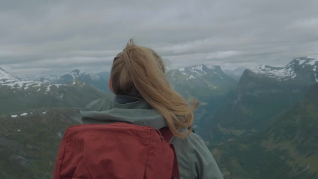 Close up of a woman facing away overlooking a vast mountainous valley with snow capped peaks wearing a jacket and backpack on an overcast day high in the Norwegian alps