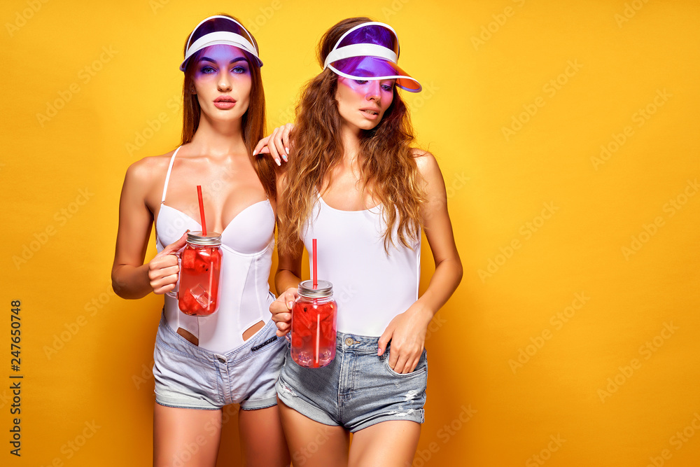 Wall mural Two sensual young women in summer outfits, cap, swimsuits holding jars with fresh beverages while standing on bright yellow background