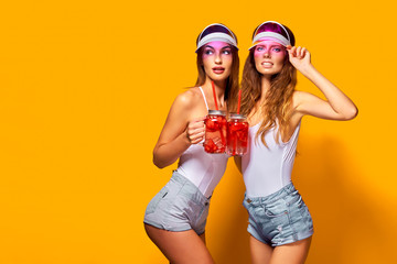 Two sensual young women in summer outfits, cap, swimsuits holding jars with fresh beverages while standing on bright yellow background