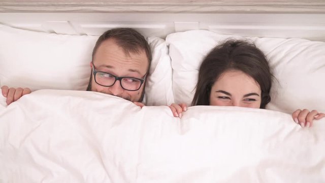 Young couple lying on the bed and looking out of the blanket. They look at the camera, at each other, blink and smile.