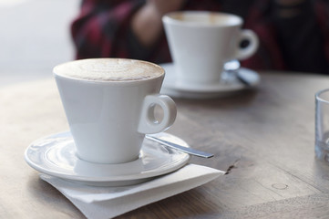 Cappuccino (coffee) in a white cup on a white saucer. In the background a blurred fragment of a man. Shallow depth of field, selective sharpness, daylight