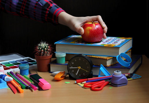 Front view of the table where school supplies, books, watches, cactus on a black background and a child's hand in a checkered shirt holds a red apple. Back to school. The concept of healthy food