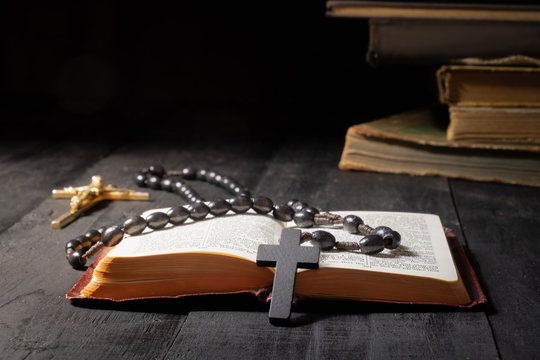 Open book of Bible and crucifix on dark table. Low-key image of New Testament, cross and rosary in bright light among darkness and shadows