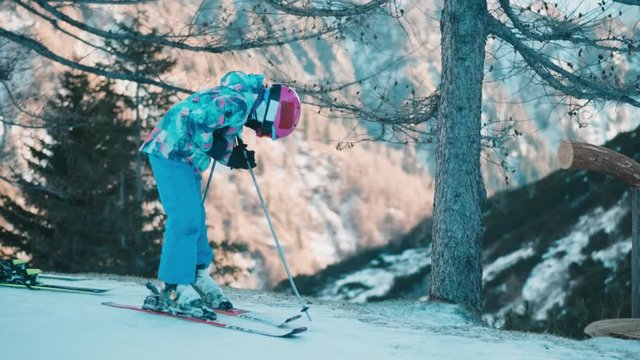 Little girl unlocking ski boot on white snowy slopes with trees & mountain in background