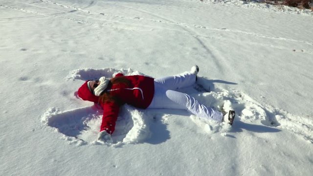 A slow motion side view of a girl making a snow angel in the snow during a bright and sunny winter afternoon outdoors.