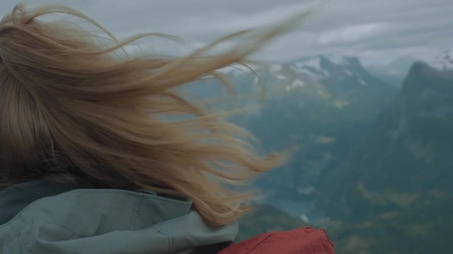 Close up of a woman facing away overlooking a vast mountainous valley with snow capped peaks wearing a jacket and backpack on an overcast day high in the Norwegian alps
