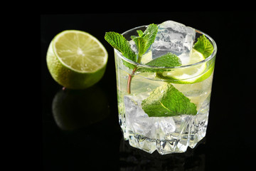 Glass of cocktail with rum, lime, ice cubes and mint leaves on black mirror background.