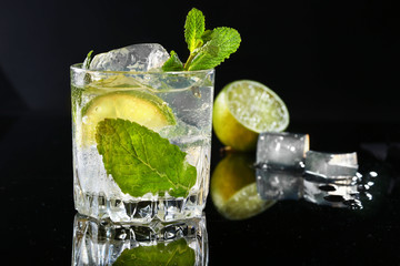Glass of cocktail with rum, lime, ice cubes and mint leaves on black mirror background.