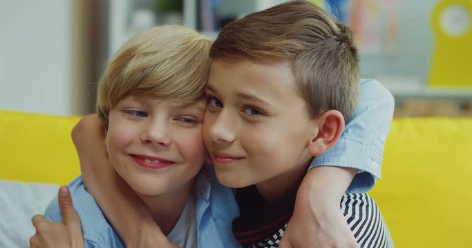 Close up of the Caucasian teen smiled boys brothers or best friends hugging while sitting on the yellow sofa in the living room. Portrait.