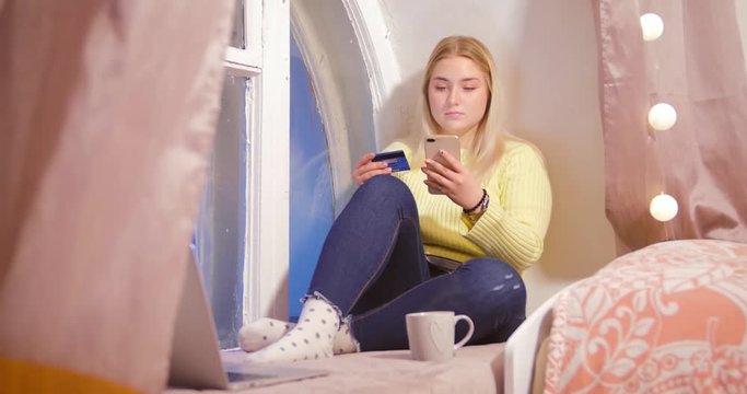 A blonde girl sits with a telephone in her room amid pink curtains and garlands and scrolls the news line on the phone in search of the right product.