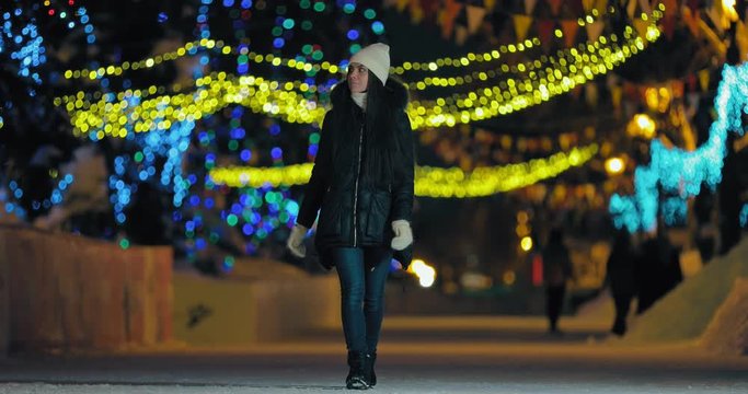 Cheerful girl walks on the night street in winter. Street is lit with garlands of lights.