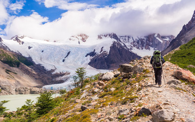 Young man walking up to mount Torre view point next to a lake or lagoon, with a green backpack on, on a summer sunny day with mountains and a glacier, near campsite De Agostini, El Chalten, Patagonia