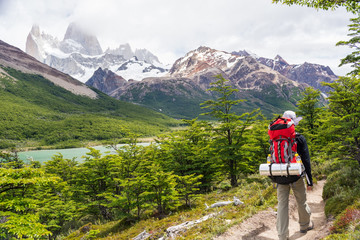 Young man walking with a red backpack on to campsite Poincenot, with mount Fitz Roy on the background on a cloudy spring day,  in El Chalten, Patagonia Argentina